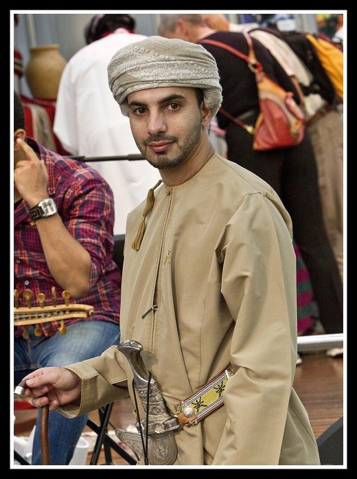 An Omani man wearing a beige dishdasha, a Musar with a Khanjar worn at his waist, holding an assa.